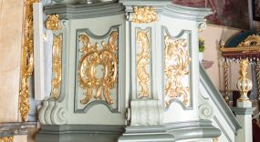 Zdjęcie nr 1: Ambona przyścienna, wisząca, z dostępem od strony prezbiterium w postaci jednobiegowych schodów z pełną balustradą dekorowaną dwoma płycinami w kształcie rombów oraz poręczą w formie gzymsu, zakończoną filarem z rokokowym wazonem. Kosz na planie kwadratu o ściętych narożach i wklęsłych bokach, na profilowanym cokole; u spodu podwieszony ostrosłup o wklęsłych bokach, z wolutami w narożach, zakończony szyszką w formie rocaille'u. Ścianki kosza flankowane pilastrami w formie spływów wolutowych z płyciną pośrodku w kształcie stojącego prostokąta o wklęsłych górnych i spodnich bokach; w polu ścianek kosza płyciny analogiczne jak na spływach wolutowych z dekoracją rocaille'ową w polu. Parapet w postaci profilowanego gzymsu. Zaplecek w kształcie stojącego prostokąta o wklęsłych bokach, ujętych spływami wolutowymi. W polu obraz Chrystusa Dobrego Pasterza umieszczony w płycinie w formie stojącego prostokąta o wklęsłych bokach, zamkniętego łukiem nadwieszonym z uskokami; u góry rocaille i rozety. Zaplecek flankowany uszami w postaci strzępionych liści. Baldachim na planie analogicznym jak kosz w postaci pełnego belkowania dekorowanego ornamentem rocaille'owym. Belkowanie stanowi podstawę dla czterech wolutowych kabłąków, dźwigających cokół z rokokowym wazonem. U spodu baldachimu rzeźbiona gołębica Ducha Świętego w promienistej glorii. Struktura polichromowana w kolorze seledynowym z ciemniejszymi profilami, ornamentyka złocona, gołębica srebrzona.