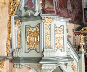 Zdjęcie nr 1: Ambona przyścienna, wisząca, z dostępem od strony prezbiterium w postaci jednobiegowych schodów z pełną balustradą dekorowaną dwoma płycinami w kształcie rombów oraz poręczą w formie gzymsu, zakończoną filarem z rokokowym wazonem. Kosz na planie kwadratu o ściętych narożach i wklęsłych bokach, na profilowanym cokole; u spodu podwieszony ostrosłup o wklęsłych bokach, z wolutami w narożach, zakończony szyszką w formie rocaille'u. Ścianki kosza flankowane pilastrami w formie spływów wolutowych z płyciną pośrodku w kształcie stojącego prostokąta o wklęsłych górnych i spodnich bokach; w polu ścianek kosza płyciny analogiczne jak na spływach wolutowych z dekoracją rocaille'ową w polu. Parapet w postaci profilowanego gzymsu. Zaplecek w kształcie stojącego prostokąta o wklęsłych bokach, ujętych spływami wolutowymi. W polu obraz Chrystusa Dobrego Pasterza umieszczony w płycinie w formie stojącego prostokąta o wklęsłych bokach, zamkniętego łukiem nadwieszonym z uskokami; u góry rocaille i rozety. Zaplecek flankowany uszami w postaci strzępionych liści. Baldachim na planie analogicznym jak kosz w postaci pełnego belkowania dekorowanego ornamentem rocaille'owym. Belkowanie stanowi podstawę dla czterech wolutowych kabłąków, dźwigających cokół z rokokowym wazonem. U spodu baldachimu rzeźbiona gołębica Ducha Świętego w promienistej glorii. Struktura polichromowana w kolorze seledynowym z ciemniejszymi profilami, ornamentyka złocona, gołębica srebrzona.