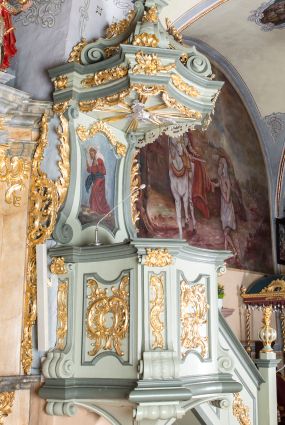 Zdjęcie nr 1: Ambona przyścienna, wisząca, z dostępem od strony prezbiterium w postaci jednobiegowych schodów z pełną balustradą dekorowaną dwoma płycinami w kształcie rombów oraz poręczą w formie gzymsu, zakończoną filarem z rokokowym wazonem. Kosz na planie kwadratu o ściętych narożach i wklęsłych bokach, na profilowanym cokole; u spodu podwieszony ostrosłup o wklęsłych bokach, z wolutami w narożach, zakończony szyszką w formie rocaille'u. Ścianki kosza flankowane pilastrami w formie spływów wolutowych z płyciną pośrodku w kształcie stojącego prostokąta o wklęsłych górnych i spodnich bokach; w polu ścianek kosza płyciny analogiczne jak na spływach wolutowych z dekoracją rocaille'ową w polu. Parapet w postaci profilowanego gzymsu. Zaplecek w kształcie stojącego prostokąta o wklęsłych bokach, ujętych spływami wolutowymi. W polu obraz Chrystusa Dobrego Pasterza umieszczony w płycinie w formie stojącego prostokąta o wklęsłych bokach, zamkniętego łukiem nadwieszonym z uskokami; u góry rocaille i rozety. Zaplecek flankowany uszami w postaci strzępionych liści. Baldachim na planie analogicznym jak kosz w postaci pełnego belkowania dekorowanego ornamentem rocaille'owym. Belkowanie stanowi podstawę dla czterech wolutowych kabłąków, dźwigających cokół z rokokowym wazonem. U spodu baldachimu rzeźbiona gołębica Ducha Świętego w promienistej glorii. Struktura polichromowana w kolorze seledynowym z ciemniejszymi profilami, ornamentyka złocona, gołębica srebrzona.