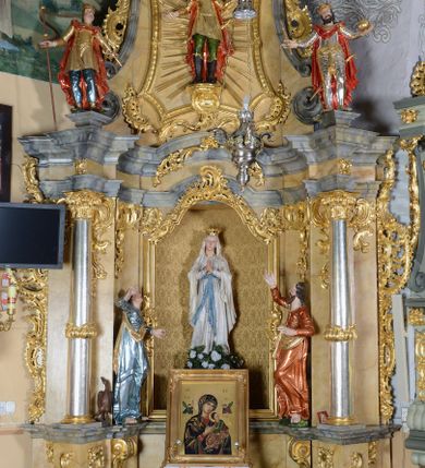 Zdjęcie nr 1: Nastawa ołtarzowa architektoniczna, jednoosiowa, jednokondygnacyjna ze zwieńczeniem. Ołtarz drewniany, prostopadłościenny, mensa drewniana z kamiennym portatylem. Antepedium w kształcie leżącego prostokąta, ujętego profilowaną ramą z aplikowanym monogramem Maryii w polu. Na ołtarzu ustawiony obraz Matki Boskiej Nieustającej Pomocy. Retabulum ustawione na dwustrefowym cokole, ujęte parą kolumn o kapitelach kompozytowych, przewiązanych poniżej połowy rocaille&#039;ową opaską, stojących na tle zwielokrotnionych pilastrów. Podpory dźwigają pełne belkowanie, wyłamane na ich osi, ustawione ukośnie, nad polem środkowym przyjmujące kształt łuku odcinkowego. W polu środkowym głęboka nisza w formie stojącego prostokąta zamkniętego łukiem wklęsło-wypukłym z uskokami, ujęta profilowaną ramą, z figurą Matki Boskiej z Lourdes. Niszę flankują dwie figury: św. Jana i św. Mateusza ustawione na konsolach. Na belkowaniu, na linii podpór cokoły z figurami św. Emeryka i św. Władysława. Zwieńczenie przyjmuje kształt stojącego prostokąta flankowanego spływami wolutowymi, zamkniętego gzymsem przechodzącym w dwie woluty. Całość zakończona promienistą glorią z monogramem Marii otoczonym obłokami, flankowana figurkami aniołków. W polu zwieńczenia rama w formie stojącego prostokąta o wklęsłych bokach i dolnych narożach, zamknięta wolutami, z figurą św. Stefana w polu, ustawioną na wsporniku. Nastawa ujęta po bokach ażurowymi uszami utworzonymi z rocaille&#039;u i prostej karbowanej wstęgi. Całość obficie dekorowana ornamentem rocaille&#039;owym oraz zwisami kwiatowymi. Struktura marmoryzowana w kolorze żółtym, profile marmoryzowane na seledynowo, ornamentyka i profilowania złocone.

