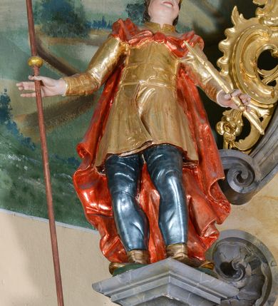 Zdjęcie nr 2: Rzeźba ścięta z tyłu, drążona, przedstawia św. Emeryka. Figura ustawiona frontalnie, w delikatnym kontrapoście, z głową skierowaną ku górze. Ręce rozłożone na boki, w lewej dłoni trzyma berło, w prawej laskę. Twarz szeroka, o pełnych, wyolbrzymionych policzkach, bez zarostu, usta delikatnie rozchylone, nos długi i szeroki. Włosy krótkie, kręcone, zasłaniające uszy, na głowie zamknięta korona. Święty ubrany jest w brązowe buty, niebieskie spodnie, złocony kaftan z dekoracyjnym pasem w talii oraz czerwony płaszcz zapięty fibulą na piersi i opadający na plecy. Polichromia naturalistyczna w odsłoniętych partiach ciała, strój i atrybuty złocone.