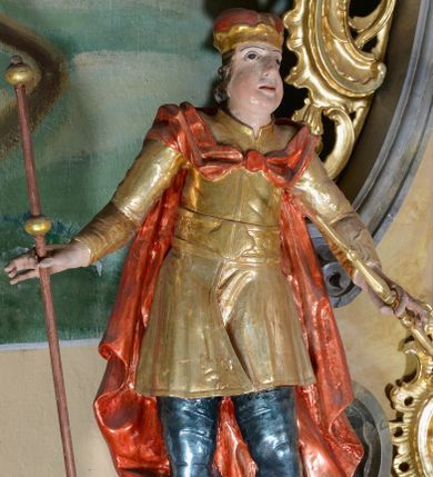 Zdjęcie nr 1: Rzeźba ścięta z tyłu, drążona, przedstawia św. Emeryka. Figura ustawiona frontalnie, w delikatnym kontrapoście, z głową skierowaną ku górze. Ręce rozłożone na boki, w lewej dłoni trzyma berło, w prawej laskę. Twarz szeroka, o pełnych, wyolbrzymionych policzkach, bez zarostu, usta delikatnie rozchylone, nos długi i szeroki. Włosy krótkie, kręcone, zasłaniające uszy, na głowie zamknięta korona. Święty ubrany jest w brązowe buty, niebieskie spodnie, złocony kaftan z dekoracyjnym pasem w talii oraz czerwony płaszcz zapięty fibulą na piersi i opadający na plecy. Polichromia naturalistyczna w odsłoniętych partiach ciała, strój i atrybuty złocone.