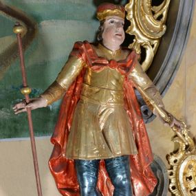 Zdjęcie nr 1: Rzeźba ścięta z tyłu, drążona, przedstawia św. Emeryka. Figura ustawiona frontalnie, w delikatnym kontrapoście, z głową skierowaną ku górze. Ręce rozłożone na boki, w lewej dłoni trzyma berło, w prawej laskę. Twarz szeroka, o pełnych, wyolbrzymionych policzkach, bez zarostu, usta delikatnie rozchylone, nos długi i szeroki. Włosy krótkie, kręcone, zasłaniające uszy, na głowie zamknięta korona. Święty ubrany jest w brązowe buty, niebieskie spodnie, złocony kaftan z dekoracyjnym pasem w talii oraz czerwony płaszcz zapięty fibulą na piersi i opadający na plecy. Polichromia naturalistyczna w odsłoniętych partiach ciała, strój i atrybuty złocone.