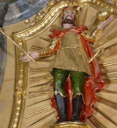 Zdjęcie nr 1: Rzeźba ścięta z tyłu, drążona, przedstawia św. Stefana. Figura ustawiona frontalnie, w delikatnym kontrapoście, z głową skierowaną ku górze. Ręce rozłożone na boki, w lewej dłoni trzyma jabłko królewskie, w prawej berło. Twarz pociągła, szczupła, okolona krótkim zarostem, z rozchylonymi ustami. Włosy krótkie, kręcone, na głowie zamknięta korona. Święty jest ubrany w buty z wysokimi cholewami, zielone spodnie oraz złocony kaftan z dekoracyjnym pasem w talii oraz czerwony płaszcz spięty na piersi broszą. Z tyłu ma zawieszony miecz, którego rękojeść jest wyrzeźbiona w kształcie głowy ptaka. Polichromia naturalistyczna w odsłoniętych partiach ciała, strój i atrybuty złocone.