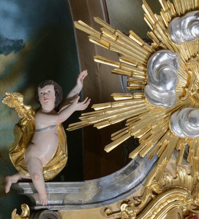 Zdjęcie nr 1: Figury dwóch aniołów adorujące monogram Marii umieszczony na tle promienistej glorii w zwieńczeniu ołtarza. Aniołki o silnie wygiętych sylwetkach z rękami wzniesionymi ku górze w stronę glorii. Sylwetki pulchne z jednym złoconym skrzydłem, umieszczonym od strony zewnętrznej ołtarza. Twarze owalne, z małymi i delikatnie rozchylonymi ustami, krótkimi i szerokimi noskami, oczami skierowanymi na wprost. Włosy ciemnobrązowe, średniej długości, zasłaniające uszy, falowane. Anioły przewiązane wąską tkaniną, odsłaniającą z jednej strony nagie biodro, anioł po lewej z tkaniną przewieszoną na srebrnym pasku. Polichromia w odsłoniętych partiach ciała naturalistyczna, szaty i skrzydła złocone.