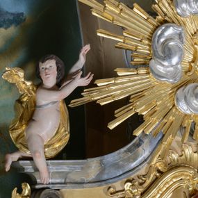 Zdjęcie nr 1: Figury dwóch aniołów adorujące monogram Marii umieszczony na tle promienistej glorii w zwieńczeniu ołtarza. Aniołki o silnie wygiętych sylwetkach z rękami wzniesionymi ku górze w stronę glorii. Sylwetki pulchne z jednym złoconym skrzydłem, umieszczonym od strony zewnętrznej ołtarza. Twarze owalne, z małymi i delikatnie rozchylonymi ustami, krótkimi i szerokimi noskami, oczami skierowanymi na wprost. Włosy ciemnobrązowe, średniej długości, zasłaniające uszy, falowane. Anioły przewiązane wąską tkaniną, odsłaniającą z jednej strony nagie biodro, anioł po lewej z tkaniną przewieszoną na srebrnym pasku. Polichromia w odsłoniętych partiach ciała naturalistyczna, szaty i skrzydła złocone.