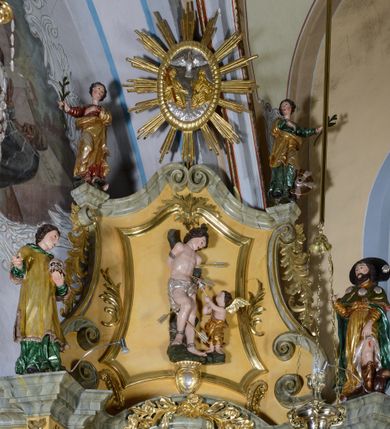 Zdjęcie nr 5: Nastawa ołtarzowa architektoniczna, jednoosiowa, jednokondygnacyjna ze zwieńczeniem. Ołtarz drewniany, prostopadłościenny, mensa drewniana z kamiennym portatylem. Antepedium w kształcie leżącego prostokąta, ujętego profilowaną ramą. Na ołtarzu tabernakulum w formie prostopadłościennej skrzynki, ujętej w narożach pilastrami o ornamentalnych trzonach, dźwigających pełne belkowanie opasujące tabernakulum z trzech stron, wyłamane na ich osi. Drzwiczki w formie stojącego prostokąta zamkniętego łukiem nadwieszonym. Retabulum ustawione na dwustrefowym cokole, ujęte parą kolumn o kapitelach kompozytowych, stojących na tle zwielokrotnionych pilastrów. Podpory dźwigają pełne belkowanie, wyłamane na ich osi, ustawione ukośnie, nad polem środkowym przyjmujące kształt łuku odcinkowego. W polu środkowym głęboka nisza w formie stojącego prostokąta zamkniętego łukiem wklęsło-wypukłym z uskokami, ujęta profilowaną ramą, z figurą Najświętszego Serca Jezusa. Niszę flankują dwie figury: św. Marka i św. Łukasza ustawione na konsolach. Na belkowaniu, na linii podpór cokoły z figurami św. Alojzego Gonzagi i św. Rocha. Zwieńczenie przyjmuje kształt stojącego prostokąta flankowanego spływami wolutowymi, zamkniętego gzymsem przechodzącym w dwie woluty. Całość zakończona owalnym medalionem z płaskorzeźbionym przedstawieniem Trójcy Świętej w promienistej glorii, flankowana figurkami św. Katarzyny Aleksandryjskiej i nierozpoznanej świętej. W polu zwieńczenia rama w formie stojącego prostokąta o wklęsłych bokach i dolnych narożach, zamknięta wolutami z akroterionem, z figurą św. Sebastiana w polu, ustawioną na wsporniku. Nastawa ujęta po bokach ażurowymi uszami utworzonymi z akantu i prostej karbowanej wstęgi. Całość obficie dekorowana ornamentem rocaille'owym oraz zwisami kwiatowymi. Struktura marmoryzowana w kolorze żółtym, profile marmoryzowane na seledynowo, ornamentyka i profilowania złocone.