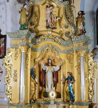 Zdjęcie nr 1: Nastawa ołtarzowa architektoniczna, jednoosiowa, jednokondygnacyjna ze zwieńczeniem. Ołtarz drewniany, prostopadłościenny, mensa drewniana z kamiennym portatylem. Antepedium w kształcie leżącego prostokąta, ujętego profilowaną ramą. Na ołtarzu tabernakulum w formie prostopadłościennej skrzynki, ujętej w narożach pilastrami o ornamentalnych trzonach, dźwigających pełne belkowanie opasujące tabernakulum z trzech stron, wyłamane na ich osi. Drzwiczki w formie stojącego prostokąta zamkniętego łukiem nadwieszonym. Retabulum ustawione na dwustrefowym cokole, ujęte parą kolumn o kapitelach kompozytowych, stojących na tle zwielokrotnionych pilastrów. Podpory dźwigają pełne belkowanie, wyłamane na ich osi, ustawione ukośnie, nad polem środkowym przyjmujące kształt łuku odcinkowego. W polu środkowym głęboka nisza w formie stojącego prostokąta zamkniętego łukiem wklęsło-wypukłym z uskokami, ujęta profilowaną ramą, z figurą Najświętszego Serca Jezusa. Niszę flankują dwie figury: św. Marka i św. Łukasza ustawione na konsolach. Na belkowaniu, na linii podpór cokoły z figurami św. Alojzego Gonzagi i św. Rocha. Zwieńczenie przyjmuje kształt stojącego prostokąta flankowanego spływami wolutowymi, zamkniętego gzymsem przechodzącym w dwie woluty. Całość zakończona owalnym medalionem z płaskorzeźbionym przedstawieniem Trójcy Świętej w promienistej glorii, flankowana figurkami św. Katarzyny Aleksandryjskiej i nierozpoznanej świętej. W polu zwieńczenia rama w formie stojącego prostokąta o wklęsłych bokach i dolnych narożach, zamknięta wolutami z akroterionem, z figurą św. Sebastiana w polu, ustawioną na wsporniku. Nastawa ujęta po bokach ażurowymi uszami utworzonymi z akantu i prostej karbowanej wstęgi. Całość obficie dekorowana ornamentem rocaille'owym oraz zwisami kwiatowymi. Struktura marmoryzowana w kolorze żółtym, profile marmoryzowane na seledynowo, ornamentyka i profilowania złocone.