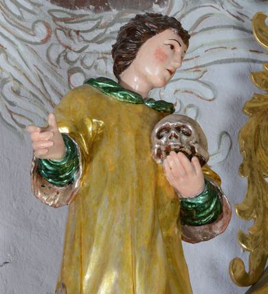 Zdjęcie nr 1: Rzeźba przedstawiająca św. Alojzego Gonzagę, pełnoplastyczna. Święty ustawiony w delikatnym kontrapoście z głową zwróconą w swoją lewą stroną, z prawą ręką zgiętą w łokciu i wysuniętą do przodu, w lewej dłoni trzyma czaszkę. Twarz szeroka z małymi i delikatnie otwartymi ustami, z prostym nosem, z podłużnymi oczami okolonymi wąskimi łukami brwiowymi, policzki silnie zarumienione. Włosy średniej długości, ciemnobrązowe, zaczesane do tyłu w bujne loki. Święty jest ubrany w długą, zieloną szatę z długimi rękawami oraz złoconą komżę, na stopach ma czarne buty. Polichromia w odsłoniętych partiach ciała naturalistyczna, jasna.