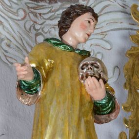 Zdjęcie nr 1: Rzeźba przedstawiająca św. Alojzego Gonzagę, pełnoplastyczna. Święty ustawiony w delikatnym kontrapoście z głową zwróconą w swoją lewą stroną, z prawą ręką zgiętą w łokciu i wysuniętą do przodu, w lewej dłoni trzyma czaszkę. Twarz szeroka z małymi i delikatnie otwartymi ustami, z prostym nosem, z podłużnymi oczami okolonymi wąskimi łukami brwiowymi, policzki silnie zarumienione. Włosy średniej długości, ciemnobrązowe, zaczesane do tyłu w bujne loki. Święty jest ubrany w długą, zieloną szatę z długimi rękawami oraz złoconą komżę, na stopach ma czarne buty. Polichromia w odsłoniętych partiach ciała naturalistyczna, jasna.