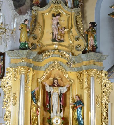 Zdjęcie nr 3: Rzeźba pełnoplastyczna przedstawia św. Sebastiana w asyście aniołka. Święty przywiązany do pnia w postawie stojącej z prawą nogą ugiętą w kolanie, z rękami i nogami skrępowanymi sznurami, głowa przechylona w jego lewą stronę. Twarz o młodzieńczych rysach, szeroka, usta drobne i otwarte, nos krótki i prosty, oczy podłużne okolone delikatną linią brwiową. Włosy ciemnobrązowe, średniej długości, zasłaniające uszy, zaczesane do tyłu bujnymi lokami. Ciało owinięte w biodrach jasną przepaską. Święty ugodzony czterema strzałami: dwiema w lewy bok i dwiema  w prawą nogę. Po lewej stronie świętego aniołek ze złotymi skrzydłami, sięgający do wysokości bioder świętego, ustawiony w pozycji stojącej, ukazany z lewego profilu, skierowany w stronę głównej postaci. W uniesionych rękach trzyma strzałę. Anioł przepasany jest w biodrach złotą szatą sięgającą za kolana, miękko drapowaną. Polichromia w odsłoniętych partiach ciała naturalistyczna, szaty i detale złocone i srebrzone. 
