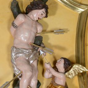 Zdjęcie nr 1: Rzeźba pełnoplastyczna przedstawia św. Sebastiana w asyście aniołka. Święty przywiązany do pnia w postawie stojącej z prawą nogą ugiętą w kolanie, z rękami i nogami skrępowanymi sznurami, głowa przechylona w jego lewą stronę. Twarz o młodzieńczych rysach, szeroka, usta drobne i otwarte, nos krótki i prosty, oczy podłużne okolone delikatną linią brwiową. Włosy ciemnobrązowe, średniej długości, zasłaniające uszy, zaczesane do tyłu bujnymi lokami. Ciało owinięte w biodrach jasną przepaską. Święty ugodzony czterema strzałami: dwiema w lewy bok i dwiema  w prawą nogę. Po lewej stronie świętego aniołek ze złotymi skrzydłami, sięgający do wysokości bioder świętego, ustawiony w pozycji stojącej, ukazany z lewego profilu, skierowany w stronę głównej postaci. W uniesionych rękach trzyma strzałę. Anioł przepasany jest w biodrach złotą szatą sięgającą za kolana, miękko drapowaną. Polichromia w odsłoniętych partiach ciała naturalistyczna, szaty i detale złocone i srebrzone. 

