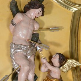 Zdjęcie nr 1: Rzeźba pełnoplastyczna przedstawia św. Sebastiana w asyście aniołka. Święty przywiązany do pnia w postawie stojącej z prawą nogą ugiętą w kolanie, z rękami i nogami skrępowanymi sznurami, głowa przechylona w jego lewą stronę. Twarz o młodzieńczych rysach, szeroka, usta drobne i otwarte, nos krótki i prosty, oczy podłużne okolone delikatną linią brwiową. Włosy ciemnobrązowe, średniej długości, zasłaniające uszy, zaczesane do tyłu bujnymi lokami. Ciało owinięte w biodrach jasną przepaską. Święty ugodzony czterema strzałami: dwiema w lewy bok i dwiema  w prawą nogę. Po lewej stronie świętego aniołek ze złotymi skrzydłami, sięgający do wysokości bioder świętego, ustawiony w pozycji stojącej, ukazany z lewego profilu, skierowany w stronę głównej postaci. W uniesionych rękach trzyma strzałę. Anioł przepasany jest w biodrach złotą szatą sięgającą za kolana, miękko drapowaną. Polichromia w odsłoniętych partiach ciała naturalistyczna, szaty i detale złocone i srebrzone. 
