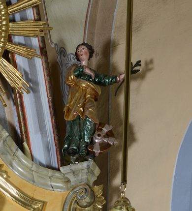 Zdjęcie nr 1: Rzeźba pełnoplastyczna przedstawiająca św. Katarzynę Aleksandryjską. Święta stoi na niskim cokole, prawą rękę ma złożoną na piersi, w lewej odsuniętej w bok trzyma palmę męczeństwa, głowę ma skierowaną w prawo. Twarz owalna o delikatnych rysach, z długim i prostym nosem, bardzo małymi ustami, okolona bujnymi włosami, zasłaniającymi uszy i podwiniętymi do tyłu w bujne fale, policzki intensywnie zarumienione. Święta jest ubrana w zieloną, długą suknię z długimi rękawami oraz złocony płaszcz założony na prawe ramię i otulający postać z przodu, na stopach ma czarne buty. Po lewej stronie świętej koło – narzędzie jej męczeństwa. Polichromia w odsłoniętych partiach ciała naturalistyczna, jasna.

