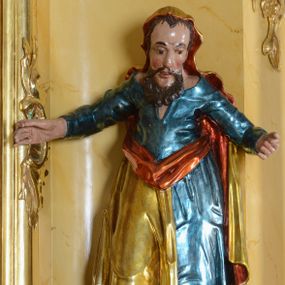 Zdjęcie nr 1: Rzeźba ścięta z tyłu, przedstawia św. Łukasza. Figura delikatnie pochylona do przodu, z obiema nogami ugiętymi w kolanach, ręce odchylone od ciała szeroko na boki, masywne dłonie. Twarz podłużna i szczupła, usta rozchylone, oczy duże, okolona średniej długości, brązową brodą. Włosy krótkie, ściśle przylegające do głowy, schowane pod płaszczem. Święty jest ubrany w niebieską suknię z długimi rękawami i  szerokim dekoltem odkrywającym ramiona oraz założony na  plecy, głowę i prawą nogę złocony płaszcz z czerwoną podszewką, stopy bose. Figura o nieanatomicznie pomniejszonej sylwetce w górnej partii. Tkanina sztywna, drapowana w grube fałdy.