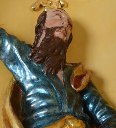 Zdjęcie nr 5: Rzeźba św. Marka ścięta z tyłu, ustawiona na niskim półokrągłym cokole. Figura ukazana w delikatnym kontrapoście z prawą nogą ugiętą w kolanie i wysuniętą do przodu, z prawą ręką uniesioną wysoko, a lewą ugiętą w łokciu i opuszczoną w dół, dłonie masywne. Święty ma głowę zwróconą ku górze i przechyloną delikatnie w lewo. Twarz podłużna, okolona długą i bujną brodą oraz średniej długości włosami opadającymi na plecy i zasłaniającymi uszy; nos długi i wąski, usta otwarte, oczy skierowane ku górze. Święty ubrany jest w niebieską suknię z długimi rękawami, przepasaną w talii oraz zarzucony na lewe ramię złoty płaszcz z czerwoną podszewką, opadający na plecy, z jedną połą upiętą na brzuchu. Tkanina sztywna, drapowana w grube fałdy. U stóp świętego głowa lwa. Polichromia w odsłoniętych partiach ciała naturalistyczna, płaszcz złocony.