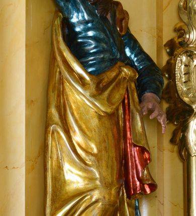 Zdjęcie nr 2: Rzeźba św. Marka ścięta z tyłu, ustawiona na niskim półokrągłym cokole. Figura ukazana w delikatnym kontrapoście z prawą nogą ugiętą w kolanie i wysuniętą do przodu, z prawą ręką uniesioną wysoko, a lewą ugiętą w łokciu i opuszczoną w dół, dłonie masywne. Święty ma głowę zwróconą ku górze i przechyloną delikatnie w lewo. Twarz podłużna, okolona długą i bujną brodą oraz średniej długości włosami opadającymi na plecy i zasłaniającymi uszy; nos długi i wąski, usta otwarte, oczy skierowane ku górze. Święty ubrany jest w niebieską suknię z długimi rękawami, przepasaną w talii oraz zarzucony na lewe ramię złoty płaszcz z czerwoną podszewką, opadający na plecy, z jedną połą upiętą na brzuchu. Tkanina sztywna, drapowana w grube fałdy. U stóp świętego głowa lwa. Polichromia w odsłoniętych partiach ciała naturalistyczna, płaszcz złocony.