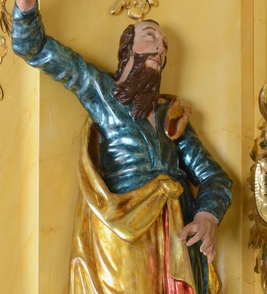Zdjęcie nr 1: Rzeźba św. Marka ścięta z tyłu, ustawiona na niskim półokrągłym cokole. Figura ukazana w delikatnym kontrapoście z prawą nogą ugiętą w kolanie i wysuniętą do przodu, z prawą ręką uniesioną wysoko, a lewą ugiętą w łokciu i opuszczoną w dół, dłonie masywne. Święty ma głowę zwróconą ku górze i przechyloną delikatnie w lewo. Twarz podłużna, okolona długą i bujną brodą oraz średniej długości włosami opadającymi na plecy i zasłaniającymi uszy; nos długi i wąski, usta otwarte, oczy skierowane ku górze. Święty ubrany jest w niebieską suknię z długimi rękawami, przepasaną w talii oraz zarzucony na lewe ramię złoty płaszcz z czerwoną podszewką, opadający na plecy, z jedną połą upiętą na brzuchu. Tkanina sztywna, drapowana w grube fałdy. U stóp świętego głowa lwa. Polichromia w odsłoniętych partiach ciała naturalistyczna, płaszcz złocony.