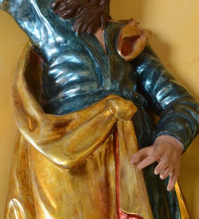 Zdjęcie nr 4: Rzeźba św. Marka ścięta z tyłu, ustawiona na niskim półokrągłym cokole. Figura ukazana w delikatnym kontrapoście z prawą nogą ugiętą w kolanie i wysuniętą do przodu, z prawą ręką uniesioną wysoko, a lewą ugiętą w łokciu i opuszczoną w dół, dłonie masywne. Święty ma głowę zwróconą ku górze i przechyloną delikatnie w lewo. Twarz podłużna, okolona długą i bujną brodą oraz średniej długości włosami opadającymi na plecy i zasłaniającymi uszy; nos długi i wąski, usta otwarte, oczy skierowane ku górze. Święty ubrany jest w niebieską suknię z długimi rękawami, przepasaną w talii oraz zarzucony na lewe ramię złoty płaszcz z czerwoną podszewką, opadający na plecy, z jedną połą upiętą na brzuchu. Tkanina sztywna, drapowana w grube fałdy. U stóp świętego głowa lwa. Polichromia w odsłoniętych partiach ciała naturalistyczna, płaszcz złocony.