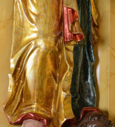 Zdjęcie nr 7: Rzeźba św. Marka ścięta z tyłu, ustawiona na niskim półokrągłym cokole. Figura ukazana w delikatnym kontrapoście z prawą nogą ugiętą w kolanie i wysuniętą do przodu, z prawą ręką uniesioną wysoko, a lewą ugiętą w łokciu i opuszczoną w dół, dłonie masywne. Święty ma głowę zwróconą ku górze i przechyloną delikatnie w lewo. Twarz podłużna, okolona długą i bujną brodą oraz średniej długości włosami opadającymi na plecy i zasłaniającymi uszy; nos długi i wąski, usta otwarte, oczy skierowane ku górze. Święty ubrany jest w niebieską suknię z długimi rękawami, przepasaną w talii oraz zarzucony na lewe ramię złoty płaszcz z czerwoną podszewką, opadający na plecy, z jedną połą upiętą na brzuchu. Tkanina sztywna, drapowana w grube fałdy. U stóp świętego głowa lwa. Polichromia w odsłoniętych partiach ciała naturalistyczna, płaszcz złocony.