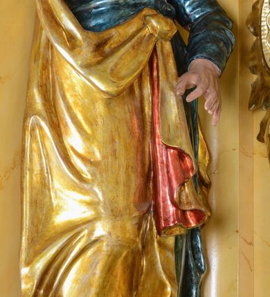 Zdjęcie nr 3: Rzeźba św. Marka ścięta z tyłu, ustawiona na niskim półokrągłym cokole. Figura ukazana w delikatnym kontrapoście z prawą nogą ugiętą w kolanie i wysuniętą do przodu, z prawą ręką uniesioną wysoko, a lewą ugiętą w łokciu i opuszczoną w dół, dłonie masywne. Święty ma głowę zwróconą ku górze i przechyloną delikatnie w lewo. Twarz podłużna, okolona długą i bujną brodą oraz średniej długości włosami opadającymi na plecy i zasłaniającymi uszy; nos długi i wąski, usta otwarte, oczy skierowane ku górze. Święty ubrany jest w niebieską suknię z długimi rękawami, przepasaną w talii oraz zarzucony na lewe ramię złoty płaszcz z czerwoną podszewką, opadający na plecy, z jedną połą upiętą na brzuchu. Tkanina sztywna, drapowana w grube fałdy. U stóp świętego głowa lwa. Polichromia w odsłoniętych partiach ciała naturalistyczna, płaszcz złocony.
