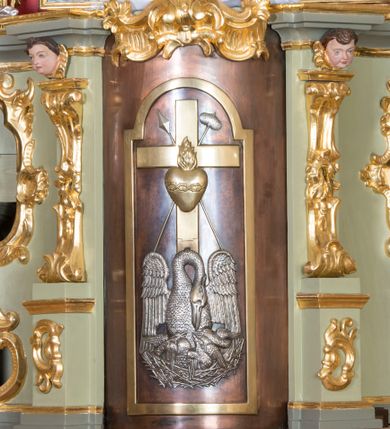 Zdjęcie nr 1: Tabernakulum w formie zbliżonej do prostopadłościennej skrzynki, ustawione na niskim cokole, o wypukłej ściance frontowej. Jest ujęte drewnianą obudową o wklęsłych bokach i flankowane parą ustawionych pod kątem pilastrów na wysokich cokołach, których trzony przyjmują formę wolut, nad nimi uskrzydlone główki aniołków. Zwieńczenie w formie profilowanego gzymsu, wyłamanego nad osiami bocznymi. Całość ujęta po bokach rocaille'owymi uszami. Część środkowa nowa, miedziana z wypukłymi drzwiczkami w formie stojącego prostokąta zamkniętego łukiem nadwieszonym. W polu aplikowane przedstawienie pelikana karmiącego młode w gnieździe, z krzyżem, gorejącym sercem i arma Christi (włócznia i gąbka z octem) powyżej. Nad drzwiczkami rocaille'owa wstęga. Tabernakulum polichromowane na kolor jasnozielony, cokół i gzyms na kolor szaro-zielony.
