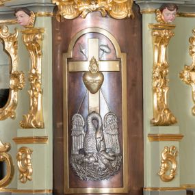 Zdjęcie nr 1: Tabernakulum w formie zbliżonej do prostopadłościennej skrzynki, ustawione na niskim cokole, o wypukłej ściance frontowej. Jest ujęte drewnianą obudową o wklęsłych bokach i flankowane parą ustawionych pod kątem pilastrów na wysokich cokołach, których trzony przyjmują formę wolut, nad nimi uskrzydlone główki aniołków. Zwieńczenie w formie profilowanego gzymsu, wyłamanego nad osiami bocznymi. Całość ujęta po bokach rocaille&#039;owymi uszami. Część środkowa nowa, miedziana z wypukłymi drzwiczkami w formie stojącego prostokąta zamkniętego łukiem nadwieszonym. W polu aplikowane przedstawienie pelikana karmiącego młode w gnieździe, z krzyżem, gorejącym sercem i arma Christi (włócznia i gąbka z octem) powyżej. Nad drzwiczkami rocaille&#039;owa wstęga. Tabernakulum polichromowane na kolor jasnozielony, cokół i gzyms na kolor szaro-zielony.
