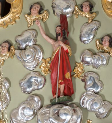 Zdjęcie nr 1: Rzeźba Chrystusa Zmartwychwstałego otoczona obłokami i uskrzydlonymi główkami anielskimi. Chrystus ukazany w postawie stojącej na niskim cokole, frontalnie, w delikatnym kontrapoście z prawą nogą ugiętą w kolanie, w lewej dłoni trzyma chorągiew, prawą wysoko uniesioną do góry czyni gest błogosławieństwa. Twarz podłużna, szczupła, z małymi i wąskimi ustami, długim i wąskim nosem, wzrokiem skierowanym na wprost, okolona długimi, ciemnobrązowymi włosami z przedziałkiem pośrodku, opadającymi na plecy oraz krótką brodą. Chrystus jest ubrany w czerwony płaszcz ze złotą podszewką, założony na lewe ramię i odsłaniający klatkę piersiową. Na dłoniach, stopach i boku widoczne ślady męki. Cokół niski, o nieregularnych bokach, pomalowany na zielono. Polichromia w odsłoniętych partiach ciała naturalistyczna, podszewka płaszcza złocona, drzewce chorągwi srebrzone. Obłoki bujne, srebrzone. W górnej części kompozycji, po bokach Chrystusa cztery uskrzydlone główki anielskie. Główki o obficie pucułowatych policzkach, wydatnych podbródkach, długich i szerokich na końcach nosach, o średniej długości ciemnobrązowych włosach z zakolami, ułożonymi po bokach w duże pukle, skrzydełka złocone.