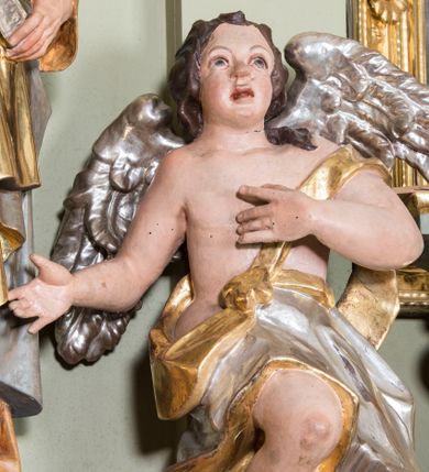 Zdjęcie nr 1: Figury aniołów flankujące tabernakulum ołtarza głównego. Anioły są ukazane w pozycji siedzącej, frontalnie, z jedną ręką złożoną na piersi, drugą odsuniętą w bok, z jedną nogą ugiętą w kolanie i widoczną spod szaty, a drugą pod nią schowaną, z głową delikatnie uniesioną ku górze; u pleców srebrne, szeroko rozłożone skrzydła. Twarz anioła po lewej owalna, o pucułowatych policzkach, mocno czerwonych i delikatnie rozchylonych ustach, małym i szerokim nosie, oczach skierowanych ku górze. Włosy ciemnobrązowe, średniej długości, kędzierzawe i zasłaniające uszy. Twarz anioła po prawej owalna, o pucułowatych policzkach, otwartych ustach, długim i szerokim na końcu nosie, oczach skierowanych do góry, okolona długimi i bujnymi włosami. Aniołek po lewej odziany jest w srebrną szatę ze złoconą podszewką, założoną na biodra i osłaniającą nogi, zawieszoną na złotym pasku przechodzącym przez piersi. Anioł po prawej ubrany jest w srebrną szatę ze złoconą podszewką założoną na prawe ramię, odsłaniającą jedną nogę. Polichromia w odsłoniętych partiach ciała naturalistyczna, szaty i skrzydła złocone i srebrzone. 