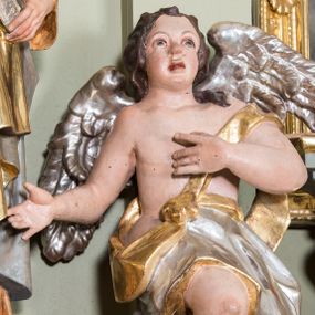 Zdjęcie nr 1: Figury aniołów flankujące tabernakulum ołtarza głównego. Anioły są ukazane w pozycji siedzącej, frontalnie, z jedną ręką złożoną na piersi, drugą odsuniętą w bok, z jedną nogą ugiętą w kolanie i widoczną spod szaty, a drugą pod nią schowaną, z głową delikatnie uniesioną ku górze; u pleców srebrne, szeroko rozłożone skrzydła. Twarz anioła po lewej owalna, o pucułowatych policzkach, mocno czerwonych i delikatnie rozchylonych ustach, małym i szerokim nosie, oczach skierowanych ku górze. Włosy ciemnobrązowe, średniej długości, kędzierzawe i zasłaniające uszy. Twarz anioła po prawej owalna, o pucułowatych policzkach, otwartych ustach, długim i szerokim na końcu nosie, oczach skierowanych do góry, okolona długimi i bujnymi włosami. Aniołek po lewej odziany jest w srebrną szatę ze złoconą podszewką, założoną na biodra i osłaniającą nogi, zawieszoną na złotym pasku przechodzącym przez piersi. Anioł po prawej ubrany jest w srebrną szatę ze złoconą podszewką założoną na prawe ramię, odsłaniającą jedną nogę. Polichromia w odsłoniętych partiach ciała naturalistyczna, szaty i skrzydła złocone i srebrzone. 