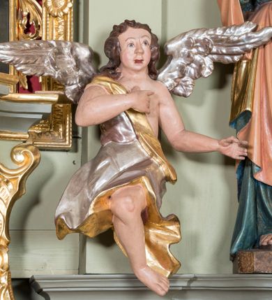 Zdjęcie nr 3: Figury aniołów flankujące tabernakulum ołtarza głównego. Anioły są ukazane w pozycji siedzącej, frontalnie, z jedną ręką złożoną na piersi, drugą odsuniętą w bok, z jedną nogą ugiętą w kolanie i widoczną spod szaty, a drugą pod nią schowaną, z głową delikatnie uniesioną ku górze; u pleców srebrne, szeroko rozłożone skrzydła. Twarz anioła po lewej owalna, o pucułowatych policzkach, mocno czerwonych i delikatnie rozchylonych ustach, małym i szerokim nosie, oczach skierowanych ku górze. Włosy ciemnobrązowe, średniej długości, kędzierzawe i zasłaniające uszy. Twarz anioła po prawej owalna, o pucułowatych policzkach, otwartych ustach, długim i szerokim na końcu nosie, oczach skierowanych do góry, okolona długimi i bujnymi włosami. Aniołek po lewej odziany jest w srebrną szatę ze złoconą podszewką, założoną na biodra i osłaniającą nogi, zawieszoną na złotym pasku przechodzącym przez piersi. Anioł po prawej ubrany jest w srebrną szatę ze złoconą podszewką założoną na prawe ramię, odsłaniającą jedną nogę. Polichromia w odsłoniętych partiach ciała naturalistyczna, szaty i skrzydła złocone i srebrzone. 