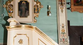 Zdjęcie nr 1: Ambona wisząca, przyścienna, złożona z kosza i schodów z balustradą, zaplecka i baldachimu ze zwieńczeniem. Kosz na planie kwadratu z wypukłą ścianą frontową, ujęty jednym pilastrem przy ścianie bocznej. Na ścianach płyciny o wykroju kwadratu z wklęsłymi narożami, wewnątrz rozety. Kosz zakończony profilowanym gzymsem, od dołu otoczony pasem z dekoracją w formie nacięć. Spięty u dołu szyszką z fryzem zdobionym czterema wolutami oraz pierścieniem z dekoracją w formie łezek. Od wschodu schody jednobiegowe z balustradą zdobioną płycinami z rozetami oraz profilowaną poręczą. Pulpit w kształcie kwadratu zamkniętego łukiem odcinkowym nadwieszonym. Zaplecek w formie stojącego prostokąta, ujęty uszami utworzonymi z ornamentu rocaille’owego połączonego z wicią i rozetami, z umieszczonymi w strukturze płaskorzeźbami postaci w niszach zamkniętych łukiem półokrągłym i wypełnionych muszlą; po lewej Jezus z barankiem, po prawej św. Kazimierz (?). Pośrodku pole obrazowe o wykroju stojącego prostokąta z wklęsłymi narożami, w nim obraz Chrystusa Dobrego Pasterza. Baldachim w formie prostokąta zamkniętego od zewnętrznej strony łukiem odcinkowym nadwieszonym; podniebie malowane na niebiesko, pośrodku pole obrazowe w formie rombu, wypełnione przedstawieniem gołębicy Ducha Świętego. Ponad baldachimem duża gloria promienista, w centrum której wizerunek Oka Opatrzności.