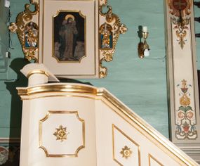 Zdjęcie nr 1: Ambona wisząca, przyścienna, złożona z kosza i schodów z balustradą, zaplecka i baldachimu ze zwieńczeniem. Kosz na planie kwadratu z wypukłą ścianą frontową, ujęty jednym pilastrem przy ścianie bocznej. Na ścianach płyciny o wykroju kwadratu z wklęsłymi narożami, wewnątrz rozety. Kosz zakończony profilowanym gzymsem, od dołu otoczony pasem z dekoracją w formie nacięć. Spięty u dołu szyszką z fryzem zdobionym czterema wolutami oraz pierścieniem z dekoracją w formie łezek. Od wschodu schody jednobiegowe z balustradą zdobioną płycinami z rozetami oraz profilowaną poręczą. Pulpit w kształcie kwadratu zamkniętego łukiem odcinkowym nadwieszonym. Zaplecek w formie stojącego prostokąta, ujęty uszami utworzonymi z ornamentu rocaille’owego połączonego z wicią i rozetami, z umieszczonymi w strukturze płaskorzeźbami postaci w niszach zamkniętych łukiem półokrągłym i wypełnionych muszlą; po lewej Jezus z barankiem, po prawej św. Kazimierz (?). Pośrodku pole obrazowe o wykroju stojącego prostokąta z wklęsłymi narożami, w nim obraz Chrystusa Dobrego Pasterza. Baldachim w formie prostokąta zamkniętego od zewnętrznej strony łukiem odcinkowym nadwieszonym; podniebie malowane na niebiesko, pośrodku pole obrazowe w formie rombu, wypełnione przedstawieniem gołębicy Ducha Świętego. Ponad baldachimem duża gloria promienista, w centrum której wizerunek Oka Opatrzności.