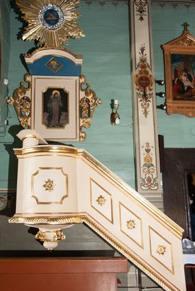 Zdjęcie nr 1: Ambona wisząca, przyścienna, złożona z kosza i schodów z balustradą, zaplecka i baldachimu ze zwieńczeniem. Kosz na planie kwadratu z wypukłą ścianą frontową, ujęty jednym pilastrem przy ścianie bocznej. Na ścianach płyciny o wykroju kwadratu z wklęsłymi narożami, wewnątrz rozety. Kosz zakończony profilowanym gzymsem, od dołu otoczony pasem z dekoracją w formie nacięć. Spięty u dołu szyszką z fryzem zdobionym czterema wolutami oraz pierścieniem z dekoracją w formie łezek. Od wschodu schody jednobiegowe z balustradą zdobioną płycinami z rozetami oraz profilowaną poręczą. Pulpit w kształcie kwadratu zamkniętego łukiem odcinkowym nadwieszonym. Zaplecek w formie stojącego prostokąta, ujęty uszami utworzonymi z ornamentu rocaille’owego połączonego z wicią i rozetami, z umieszczonymi w strukturze płaskorzeźbami postaci w niszach zamkniętych łukiem półokrągłym i wypełnionych muszlą; po lewej Jezus z barankiem, po prawej św. Kazimierz (?). Pośrodku pole obrazowe o wykroju stojącego prostokąta z wklęsłymi narożami, w nim obraz Chrystusa Dobrego Pasterza. Baldachim w formie prostokąta zamkniętego od zewnętrznej strony łukiem odcinkowym nadwieszonym; podniebie malowane na niebiesko, pośrodku pole obrazowe w formie rombu, wypełnione przedstawieniem gołębicy Ducha Świętego. Ponad baldachimem duża gloria promienista, w centrum której wizerunek Oka Opatrzności.