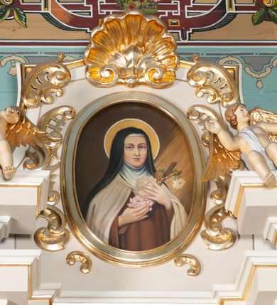 Zdjęcie nr 2: Obraz w kształcie pionowego owalu. W centrum św. Teresa ukazana frontalnie, w półpostaci, z głową lekko zwróconą na lewo. W uniesionych przed sobą dłoniach trzyma białe i jasnoróżowe róże oraz niewielki krucyfiks. Ubrana jest w strój karmelitański: brązowy habit i jasny płaszcz, białą tokę oraz czarny welon. Jej głowę otacza nimb. Tło jest jednolite, ciemnobrązowe, z prawej strony widać padające skośnie promienie. Obraz został umieszczony w profilowanej, drewnianej, złoconej ramie w zwieńczeniu ołtarza bocznego.