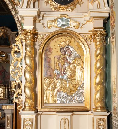 Zdjęcie nr 2: Obraz w kształcie stojącego prostokąta zamkniętego od góry łukiem półokrągłym z przedstawieniem Matki Boskiej Różańcowej. Większość kompozycji zajmuje Matka Boska tronująca na srebrnych chmurach, podtrzymująca obiema rękami Dzieciątko Jezus. Maria ma owalną twarz o łagodnym wyrazie, niebieskie oczy, wąski nos oraz niewielkie, czerwone usta, a na policzkach delikatne rumieńce. Ona, oraz pozostałe postacie sceny mają aplikowane sukienki. Maria jest ubrana w złotą suknię i srebrny welon, szaty marszczą się drobnymi, równoległymi fałdami, które układają się w różnych kierunkach. Dzieciątko stoi na srebrnej poduszce leżącej na kolanach Matki, obie ręce wyciąga w stronę klęczącego poniżej św. Dominika, prawą podaje mu aplikowany różaniec. Jezus ma owalną twarz z zaokrąglonymi policzkami, mały nos i różowe usta. Głowę okalają mu krótkie, kręcone, brązowe włosy. Jest ubrany w krótką, złotą sukienkę. Obie postacie mają na głowach korony zamknięte, wokół nich umieszczono 12 gwiazdek. Z lewej strony kompozycji u dołu klęczy św. Dominik. Prawą rękę wyciąga w stronę różańca, głowę unosi do góry, patrzy na scenę przed sobą. Ma długi, prosty nos, krótką brodę oraz wąsy, na głowie wyciętą tonsurę. Jest ubrany w habit, który również marszczy się drobnymi fałdami. Ponad nim, przy brzegu kompozycji są dwie uskrzydlone główki anielskie. Tło stanowią żółte obłoki. Obraz otacza złota, profilowana rama.