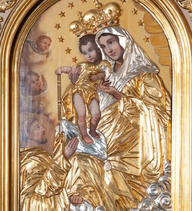 Zdjęcie nr 1: Obraz w kształcie stojącego prostokąta zamkniętego od góry łukiem półokrągłym z przedstawieniem Matki Boskiej Różańcowej. Większość kompozycji zajmuje Matka Boska tronująca na srebrnych chmurach, podtrzymująca obiema rękami Dzieciątko Jezus. Maria ma owalną twarz o łagodnym wyrazie, niebieskie oczy, wąski nos oraz niewielkie, czerwone usta, a na policzkach delikatne rumieńce. Ona, oraz pozostałe postacie sceny mają aplikowane sukienki. Maria jest ubrana w złotą suknię i srebrny welon, szaty marszczą się drobnymi, równoległymi fałdami, które układają się w różnych kierunkach. Dzieciątko stoi na srebrnej poduszce leżącej na kolanach Matki, obie ręce wyciąga w stronę klęczącego poniżej św. Dominika, prawą podaje mu aplikowany różaniec. Jezus ma owalną twarz z zaokrąglonymi policzkami, mały nos i różowe usta. Głowę okalają mu krótkie, kręcone, brązowe włosy. Jest ubrany w krótką, złotą sukienkę. Obie postacie mają na głowach korony zamknięte, wokół nich umieszczono 12 gwiazdek. Z lewej strony kompozycji u dołu klęczy św. Dominik. Prawą rękę wyciąga w stronę różańca, głowę unosi do góry, patrzy na scenę przed sobą. Ma długi, prosty nos, krótką brodę oraz wąsy, na głowie wyciętą tonsurę. Jest ubrany w habit, który również marszczy się drobnymi fałdami. Ponad nim, przy brzegu kompozycji są dwie uskrzydlone główki anielskie. Tło stanowią żółte obłoki. Obraz otacza złota, profilowana rama.