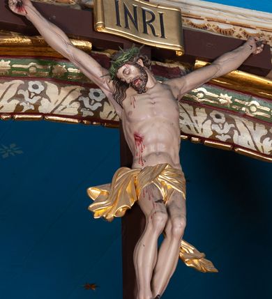 Zdjęcie nr 1: Krucyfiks, to przedstawienie ukrzyżowanego Jezusa. Martwe ciało Zbawiciela o wyraźnie zaznaczonej muskulaturze jest rozciągnięte i przybite trzema gwoździami do prostego krzyża. Widać na nim ślady męki: strużki krwi płynące po ciele, przebity bok oraz krwawiące rany. Głowa jest opuszczona na prawe ramię, oczy i usta zamknięte; na głowie zielona korona cierniowa. Chrystus ma rozpostarte ręce, stopy założone na siebie, prawa na lewej, przybite jednym gwoździem. Biodra otacza złote perizonium przewiązane z przodu z końcami rozwiewającymi się na boki. Na górnym ramieniu krzyża widnieje rozwinięty titulus z majuskułowymi literami „INRI” (łac. Iesus Nazarenus Rex Iudaeorum).