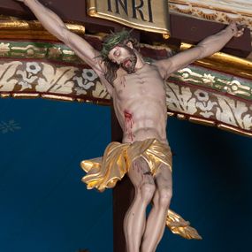 Zdjęcie nr 1: Krucyfiks, to przedstawienie ukrzyżowanego Jezusa. Martwe ciało Zbawiciela o wyraźnie zaznaczonej muskulaturze jest rozciągnięte i przybite trzema gwoździami do prostego krzyża. Widać na nim ślady męki: strużki krwi płynące po ciele, przebity bok oraz krwawiące rany. Głowa jest opuszczona na prawe ramię, oczy i usta zamknięte; na głowie zielona korona cierniowa. Chrystus ma rozpostarte ręce, stopy założone na siebie, prawa na lewej, przybite jednym gwoździem. Biodra otacza złote perizonium przewiązane z przodu z końcami rozwiewającymi się na boki. Na górnym ramieniu krzyża widnieje rozwinięty titulus z majuskułowymi literami „INRI” (łac. Iesus Nazarenus Rex Iudaeorum).