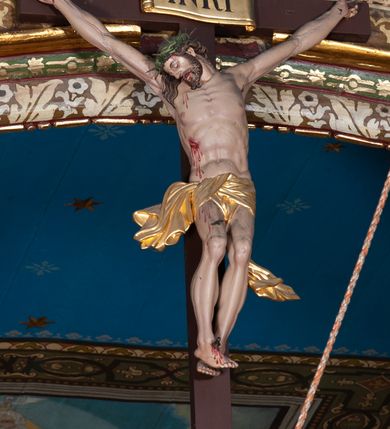 Zdjęcie nr 2: Krucyfiks, to przedstawienie ukrzyżowanego Jezusa. Martwe ciało Zbawiciela o wyraźnie zaznaczonej muskulaturze jest rozciągnięte i przybite trzema gwoździami do prostego krzyża. Widać na nim ślady męki: strużki krwi płynące po ciele, przebity bok oraz krwawiące rany. Głowa jest opuszczona na prawe ramię, oczy i usta zamknięte; na głowie zielona korona cierniowa. Chrystus ma rozpostarte ręce, stopy założone na siebie, prawa na lewej, przybite jednym gwoździem. Biodra otacza złote perizonium przewiązane z przodu z końcami rozwiewającymi się na boki. Na górnym ramieniu krzyża widnieje rozwinięty titulus z majuskułowymi literami „INRI” (łac. Iesus Nazarenus Rex Iudaeorum).