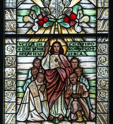 Zdjęcie nr 1: Witraż o wykroju prostokąta stojącego zakończonego łukiem odcinkowym nadwieszonym. Wewnątrz przedstawienie Chrystusa w otoczeniu sześciu postaci męskich. Chrystus w nimbie stojący frontalnie z prawą dłonią wyciągniętą w geście błogosławieństwa, lewą wskazuje na gorejące serce na piersi. Ubrany jest w białą tunikę i czerwony płaszcz przewinięty na wysokości pasa. W otoczeniu Chrystusa po lewej strony trzy postacie duchownych, z których pierwszy ubrany jest w komżę, kapę i stułę, a po prawej trzech mężczyzn w strojach chłopskich. Pierwszy z nich ma na sobie koszulę, kamizelkę, szerokie spodnie i sukmanę. Scena otoczona bordiurą z dekoracją kwiatową. Na wysokości głowy Chrystusa inskrypcja: „VCZCIE SIĘ / +ODE MNIE / IŻEM JEST / CICHY I PO/KORNEGO+ / SERCA..&quot; Powyżej sceny katusz z inskrypcją „IHS”, otoczony wolutami i czerwonymi kwiatami, w glorii. Poniżej kartusz z inskrypcją „NAJŚWIĘTSZE / +SERCE+ / +JEZUSA+ / ZMIŁUJ SIĘ NAD NAMI” również w otoczeniu wolut. Witraż utrzymany w chłodnej kolorystyce, z przewagą szarości i zieleni. 