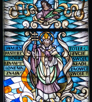 Zdjęcie nr 1: Witraż w formie stojącego prostokąta zwieńczonego łukiem odcinkowym nadwieszonym z przedstawieniem św. Wojciecha. Świętego ukazano frontalnie, całopostaciowo, ujętego stylizowanymi wstęgami i biało-niebieskimi polami romboidalnymi. Prawą rękę opiera na krzyżu patriarszym, lewą składa na piersi, patrzy przed siebie, Jest ubrany w białą albę, fioletową komżę, i zielono-żółty płaszcz. Na strój ma nałożony paliusz.  Nad głową znajduje się ujęty stylizowanymi wstęgami półpostaciowy wizerunek kobiety, która patrzy w dół i trzyma w rękach palmę męczeństwa. Jest ubrana w fioletową suknię, ma długie, opadające na plecy włosy. Wizerunek św. Wojciecha jest otoczony inskrypcją „JAM JEST / PASTERZ / DOBRY / I ZNAM SWOJE / I ONE MNIE / ZNAJĄ, JAN X 14 / TOTEŻ / I ŻYCIE / SWOJE / KŁADĘ / ZA OWCE / SWOJE JAN X 14.” U stóp świętego inskrypcja „ŚWIĘTEMV / WOJCIECHOWI / WITRAŻ TEN OFIAROWAŁA / GROMADA / POZOWICE /R(OKU) P(AŃSKIEGO) 1952.” Witraż utrzymany w chłodnej kolorystyce, z przewagą niebieskiego, zieleni i szarości. 