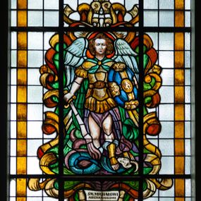 Zdjęcie nr 1: Witraż w formie stojącego prostokąta zwieńczonego łukiem trójwklęsłym. Przedstawia wizerunek św. Michała Archanioła ukazanego frontalnie, całopostaciowo, z tarczą z Alfą i Omegą w lewej ręce, ognistym mieczem w prawej i wężem pod stopami. Świety jest ubrany w strój żołnierza rzymskiego: złoty pancerz z naramiennikami założony na fioletową, rozciętą tunikę i wysokie sandały. Wizerunek ujęty zielonymi polami o nieregularnym kształcie i stylizowanymi żółto-bązowymi wstęgami. Całość przedstawienia na tle pól z niebarwionego szkła. Wzdłuż dłuższych boków witraża i łuku bordiura w postaci ciemnożółtej wstęgi. U stóp świętego inskrypcja „ŚW(IĘTEMU) MICHAŁOWI / ARCHANIOŁOWI / OFIAROWAŁA / GROMADA / POLANKA HALLER”. Witraż o intensywnych kolorach, z przewagą żółto-brązowego. 
