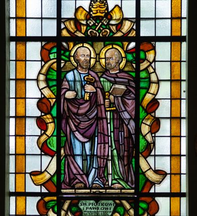 Zdjęcie nr 1: Witraż w formie stojącego prostokąta zwieńczonego łukiem trójwklęsłym. Przedstawia wizerunek św. Piotra i św. Pawła, ukazanych frontalnie, całopostaciowo. Po lewej stronie kompozycji św. Piotr z kluczem w lewej dłoni, prawą unosi zaciśniętą. Jest ubrany w niebiesko-zieloną suknię i bordowy płaszcz przerzucony przez ręce i lewe ramię. Po prawej św. Paweł z księgą w lewej i opartym o ziemię mieczem w prawej ręce. Ma na sobie zieloną suknię i bordowy płaszcz owinięty wokół szyi i lewej ręki. Powyżej świętych tiara papieska ze skrzyżowanymi poniżej kluczami. Wizerunek ujęty zielonymi polami o nieregularnym kształcie i stylizowanymi wstęgami. Całość przedstawienia na tle pól z niebarwionego szkła. Wzdłuż dłuższych boków witraża i łuku bordiura w postaci ciemnożółtej wstęgi. U stóp świętych inskrypcja „ŚW(IĘTEMU) PIOTROWI / i PAWŁOWI / WITRAŻ OFIAROWA/ŁA  GROMADA / OCHODZA / R[OKU] P[AŃSKIEGO] 1953”. Witraż o intensywnych kolorach, z przewagą bordowego i zielonego. 