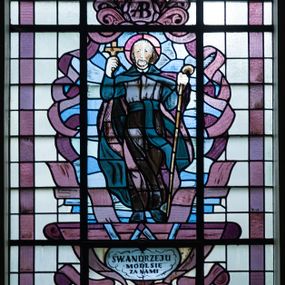 Zdjęcie nr 1: Witraż w formie stojącego prostokąta zwieńczonego łukiem trójwklęsłym. W polu wizerunek św. Andrzeja Boboli, ukazanego frontalnie, całopostaciowo, z laską pasterską w lewej dłoni i krzyżem w uniesionej prawej. Święty jest ubrany w habit oraz rozwiewający się płaszcz w odcieniach koloru bordowego i ciemnozielonego. Wizerunek ujęty blado- i ciemnoliliowymi polami o nieregularnym kształcie oraz stylizowanymi bordowymi wstęgami. Całość przedstawienia na tle pól z niebarwionego szkła. Wzdłuż dłuższych boków witraża i łuku bordiura w postaci bordowej wstęgi. U stóp świętego inskrypcja „ŚW(IĘTY) ANDRZEJU / MÓDL SIĘ / ZA NAMI” i niżej „OFIAROWAŁA / GROMADA ZELCZYNA / 1953”. Witraż o intensywnych kolorach z przewagą bordowego. 