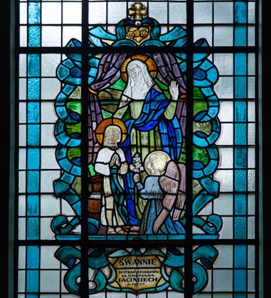 Zdjęcie nr 1: Witraż w ścianie północnej w drugim przęśle nawy bocznej, w formie stojącego prostokąta zwieńczonego łukiem trójwklęsłym. Przedstawia wizerunek św. Anny z Marią i aniołem. Święta ukazana frontalnie, wznosi w geście błogosławieństwa lewą dłoń, prawą obejmując Marię. Jest ubrana w zieloną suknię i niebieski płaszcz przerzucony przez lewe ramię, na głowie ma białą chustę.  Matka Boska, ukazana jako dziewczynka stoi i składa ręce do modlitwy. Ma na sobie białą sukienkę przewiązaną zieloną szarfą opadającą do kostek. Przed nią klęczy archanioł z lilią w lewej dłoni. Wizerunek ujęty zielonymi polami o nieregularnym kształcie oraz stylizowanymi błękitnymi wstęgami. Całość przedstawienia na tle pól z niebarwionego szkła. Wzdłuż dłuższych boków witraża i łuku bordiura w postaci błękitnej wstęgi. U stóp przedstawienia inskrypcja „ŚW(IĘTEJ) ANNIE / WITRAŻ OFIAROWA/ŁA GBROMADA / FACIMIECH / A(NNO) 1953 D(OMINI)”