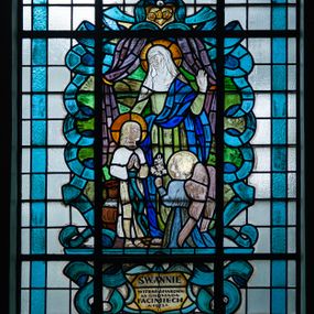 Zdjęcie nr 1: Witraż w ścianie północnej w drugim przęśle nawy bocznej, w formie stojącego prostokąta zwieńczonego łukiem trójwklęsłym. Przedstawia wizerunek św. Anny z Marią i aniołem. Święta ukazana frontalnie, wznosi w geście błogosławieństwa lewą dłoń, prawą obejmując Marię. Jest ubrana w zieloną suknię i niebieski płaszcz przerzucony przez lewe ramię, na głowie ma białą chustę.  Matka Boska, ukazana jako dziewczynka stoi i składa ręce do modlitwy. Ma na sobie białą sukienkę przewiązaną zieloną szarfą opadającą do kostek. Przed nią klęczy archanioł z lilią w lewej dłoni. Wizerunek ujęty zielonymi polami o nieregularnym kształcie oraz stylizowanymi błękitnymi wstęgami. Całość przedstawienia na tle pól z niebarwionego szkła. Wzdłuż dłuższych boków witraża i łuku bordiura w postaci błękitnej wstęgi. U stóp przedstawienia inskrypcja „ŚW(IĘTEJ) ANNIE / WITRAŻ OFIAROWA/ŁA GBROMADA / FACIMIECH / A(NNO) 1953 D(OMINI)”