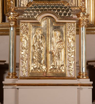 Zdjęcie nr 11: Ołtarz przyścienny, architektoniczny, na cokole, z bramkami. Nastawa jednoosiowa, jednokondygnacyjna ze zwieńczeniem. Mensa murowana, blat kamienny. Antepedium w kształcie leżącego prostokąta ujętego parami kolumn o gładkich trzonach i głowicach kompozytowych, podtrzymujących belkowanie z parami tryglifów. Belkowanie nad częścią środkową ucięte łukowato. W polu środkowym płaskorzeźbiona scena z przedstawieniem Świętej Rodziny. Postać św. Józefa półleżąca, zwrócona profilem w prawo, w stronę Matki Boskiej. Siedząca Maria skierowana w trzech czwartych w prawo, trzyma na kolanach poruszone Dzieciątko. Oś środkowa flankowana parą opilastrowanych filarów i kolumn o kapitelach kompozytowych, ustawionych przed licem ołtarza pod kątem. Podpory dźwigają pełne, wyłamane belkowanie oraz odpowiadający ich ułożeniu przerwany przyczółek. Zwieńczenie ujęte w spływy, zamknięte łukiem nadwieszonym. Cokoły kolumn dekorowane rocaille’em. W polu środkowym obraz we wnęce z przedstawieniem Matki Boskiej z Dzieciątkiem zasłaniany wizerunkiem św. Floriana w profilowanej ramie, w formie stojącego prostokąta zamkniętego dwoma, zwróconymi naprzeciwlegle stykającymi się spływami wolutowymi oraz rocaille’owym kartuszem z inskrypcją „POD TWOJĄ OBRONĘ”. Pierwsza kondygnacja ujęta uszakami utworzonymi z ornamentu rocaille’owego, ażurowej kratki regencyjnej i ornamentu roślinnego w formie kwiatów. Na bramkach ustawiono figury św. Wojciecha i św. Stanisława. Na przerwanym przyczółku spoczywa para adorujących aniołów. W zwieńczeniu profilowana rama powtarzająca jego formę z obrazem św. Antoniego Padewskiego zamknięta rocaille’em. Całość ujęta w spływy z ornamentu roślinnego. Struktura polichromowana na kremowo ze złoconą i srebrzoną ornamentyką oraz detalami architektonicznymi.