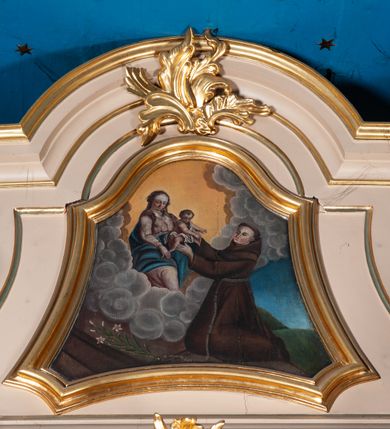 Zdjęcie nr 18: Ołtarz przyścienny, architektoniczny, na cokole, z bramkami. Nastawa jednoosiowa, jednokondygnacyjna ze zwieńczeniem. Mensa murowana, blat kamienny. Antepedium w kształcie leżącego prostokąta ujętego parami kolumn o gładkich trzonach i głowicach kompozytowych, podtrzymujących belkowanie z parami tryglifów. Belkowanie nad częścią środkową ucięte łukowato. W polu środkowym płaskorzeźbiona scena z przedstawieniem Świętej Rodziny. Postać św. Józefa półleżąca, zwrócona profilem w prawo, w stronę Matki Boskiej. Siedząca Maria skierowana w trzech czwartych w prawo, trzyma na kolanach poruszone Dzieciątko. Oś środkowa flankowana parą opilastrowanych filarów i kolumn o kapitelach kompozytowych, ustawionych przed licem ołtarza pod kątem. Podpory dźwigają pełne, wyłamane belkowanie oraz odpowiadający ich ułożeniu przerwany przyczółek. Zwieńczenie ujęte w spływy, zamknięte łukiem nadwieszonym. Cokoły kolumn dekorowane rocaille’em. W polu środkowym obraz we wnęce z przedstawieniem Matki Boskiej z Dzieciątkiem zasłaniany wizerunkiem św. Floriana w profilowanej ramie, w formie stojącego prostokąta zamkniętego dwoma, zwróconymi naprzeciwlegle stykającymi się spływami wolutowymi oraz rocaille’owym kartuszem z inskrypcją „POD TWOJĄ OBRONĘ”. Pierwsza kondygnacja ujęta uszakami utworzonymi z ornamentu rocaille’owego, ażurowej kratki regencyjnej i ornamentu roślinnego w formie kwiatów. Na bramkach ustawiono figury św. Wojciecha i św. Stanisława. Na przerwanym przyczółku spoczywa para adorujących aniołów. W zwieńczeniu profilowana rama powtarzająca jego formę z obrazem św. Antoniego Padewskiego zamknięta rocaille’em. Całość ujęta w spływy z ornamentu roślinnego. Struktura polichromowana na kremowo ze złoconą i srebrzoną ornamentyką oraz detalami architektonicznymi.