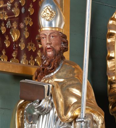 Zdjęcie nr 6: Ołtarz przyścienny, architektoniczny, na cokole, z bramkami. Nastawa jednoosiowa, jednokondygnacyjna ze zwieńczeniem. Mensa murowana, blat kamienny. Antepedium w kształcie leżącego prostokąta ujętego parami kolumn o gładkich trzonach i głowicach kompozytowych, podtrzymujących belkowanie z parami tryglifów. Belkowanie nad częścią środkową ucięte łukowato. W polu środkowym płaskorzeźbiona scena z przedstawieniem Świętej Rodziny. Postać św. Józefa półleżąca, zwrócona profilem w prawo, w stronę Matki Boskiej. Siedząca Maria skierowana w trzech czwartych w prawo, trzyma na kolanach poruszone Dzieciątko. Oś środkowa flankowana parą opilastrowanych filarów i kolumn o kapitelach kompozytowych, ustawionych przed licem ołtarza pod kątem. Podpory dźwigają pełne, wyłamane belkowanie oraz odpowiadający ich ułożeniu przerwany przyczółek. Zwieńczenie ujęte w spływy, zamknięte łukiem nadwieszonym. Cokoły kolumn dekorowane rocaille’em. W polu środkowym obraz we wnęce z przedstawieniem Matki Boskiej z Dzieciątkiem zasłaniany wizerunkiem św. Floriana w profilowanej ramie, w formie stojącego prostokąta zamkniętego dwoma, zwróconymi naprzeciwlegle stykającymi się spływami wolutowymi oraz rocaille’owym kartuszem z inskrypcją „POD TWOJĄ OBRONĘ”. Pierwsza kondygnacja ujęta uszakami utworzonymi z ornamentu rocaille’owego, ażurowej kratki regencyjnej i ornamentu roślinnego w formie kwiatów. Na bramkach ustawiono figury św. Wojciecha i św. Stanisława. Na przerwanym przyczółku spoczywa para adorujących aniołów. W zwieńczeniu profilowana rama powtarzająca jego formę z obrazem św. Antoniego Padewskiego zamknięta rocaille’em. Całość ujęta w spływy z ornamentu roślinnego. Struktura polichromowana na kremowo ze złoconą i srebrzoną ornamentyką oraz detalami architektonicznymi.